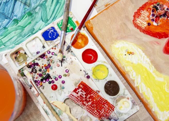 Mākslinieka darba virsma no augšas: krāsu palete ar košām krāsām, otas, krelles un dažādi fakturēti materiāli, blakus audekls ar biezu krāsu klājumu dzeltenos, oranžos un sarkanos toņos.