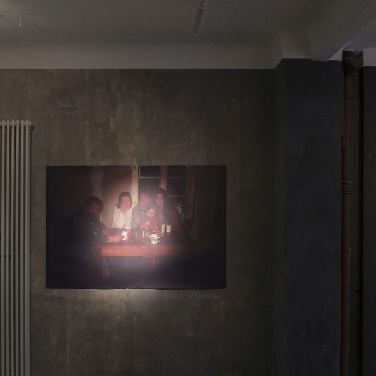 Instalācija izstāžu telpā: uz sienas pie auklas piespraustas nodilušas drēbes, centrā fotogrāfija ar cilvēkiem pie galda vājā apgaismojumā, bet labajā pusē liela jauktas tehnikas darba virsma ar tekstu un nelielu foto, industriālā, pelēkā vidē.