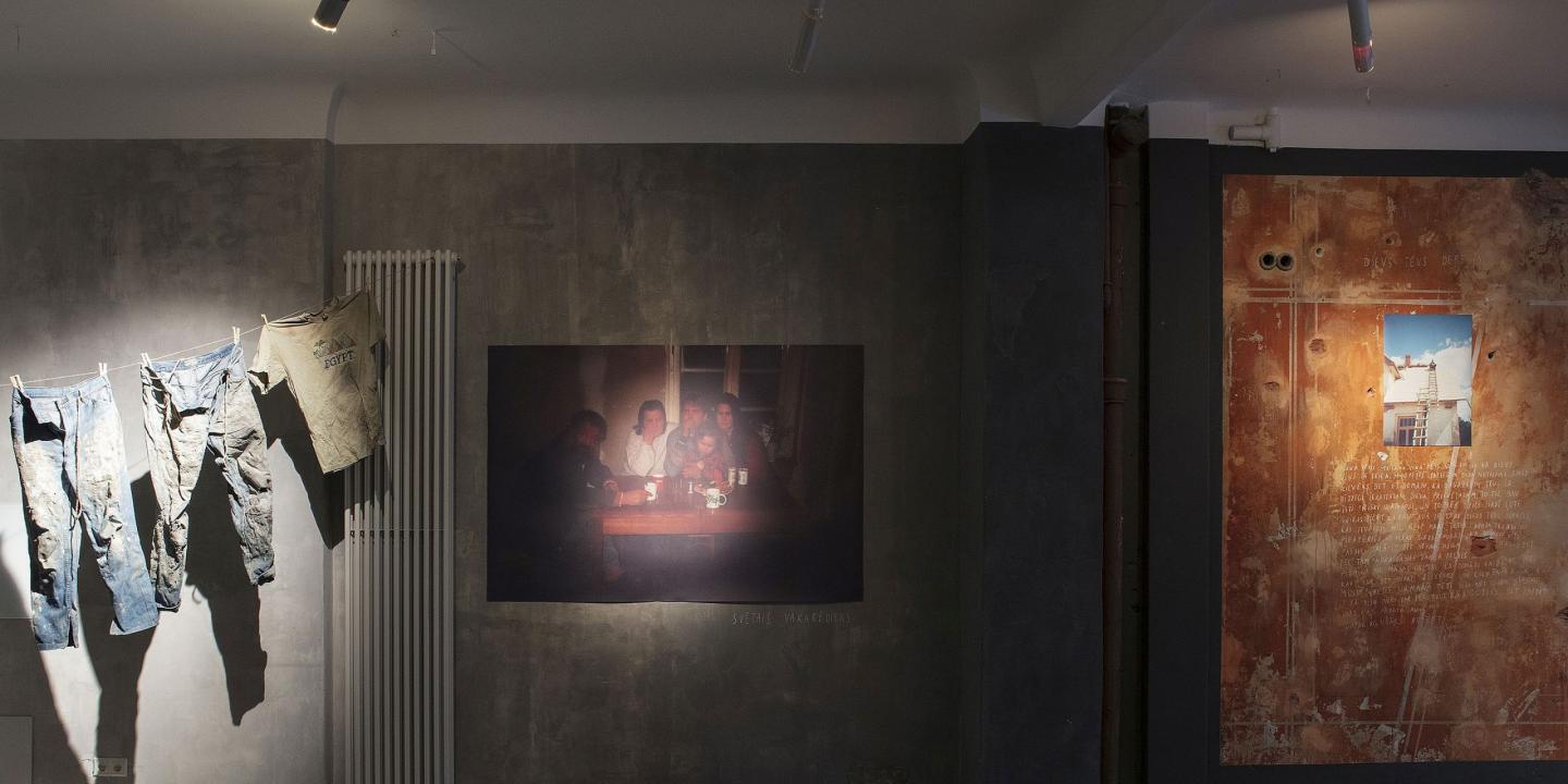 Instalācija izstāžu telpā: uz sienas pie auklas piespraustas nodilušas drēbes, centrā fotogrāfija ar cilvēkiem pie galda vājā apgaismojumā, bet labajā pusē liela jauktas tehnikas darba virsma ar tekstu un nelielu foto, industriālā, pelēkā vidē.