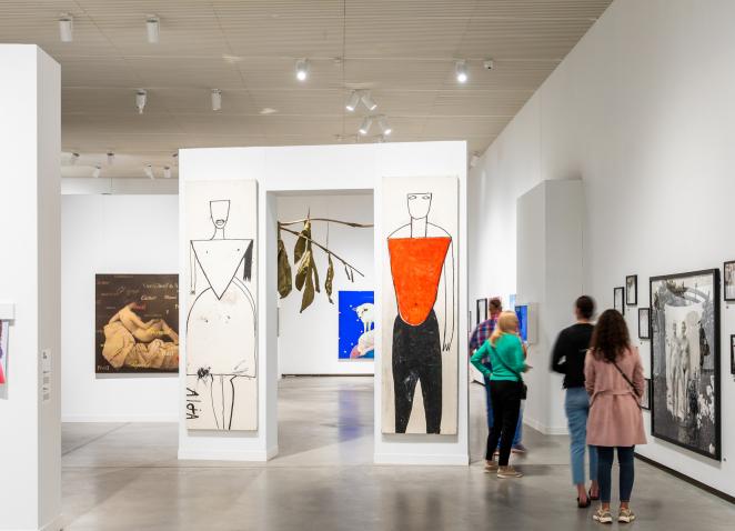 Apmeklētāji laikmetīgās mākslas izstādē aplūko gleznas un fotogrāfijas plašā, gaišā muzeja zālē.