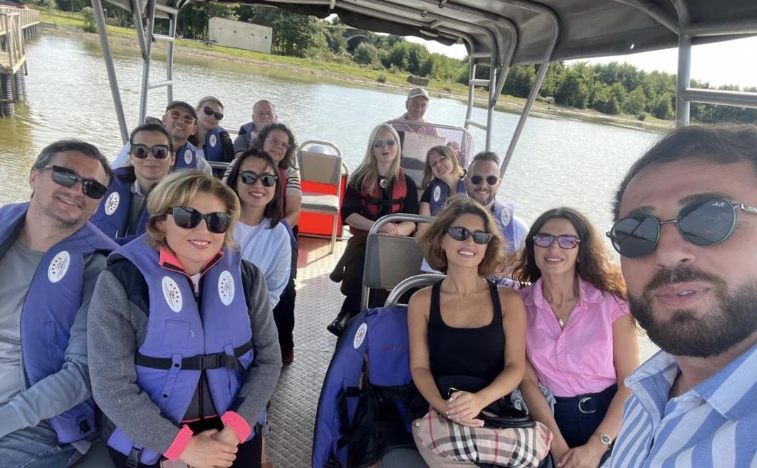 A smiling group of people wearing life jackets and sunglasses sits on the deck of a covered tour boat with a river in the background.