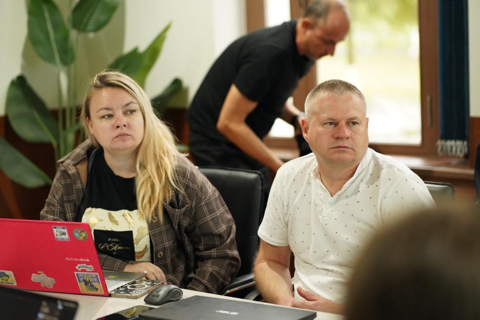 Pie galda telpā sēž sieviete pie sarkana klēpjdatora un vīrietis baltā kreklā, bet fonā stāv vēl viens vīrietis pie loga.