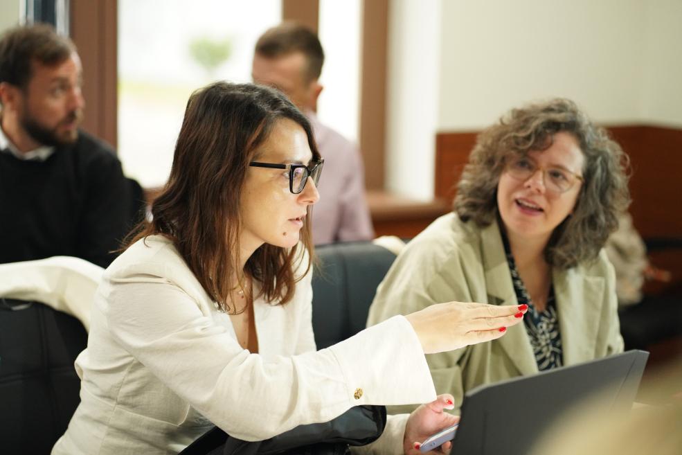 Three people sit indoors at a table, while in the foreground a woman wearing glasses and a white jacket gestures during a conversation.