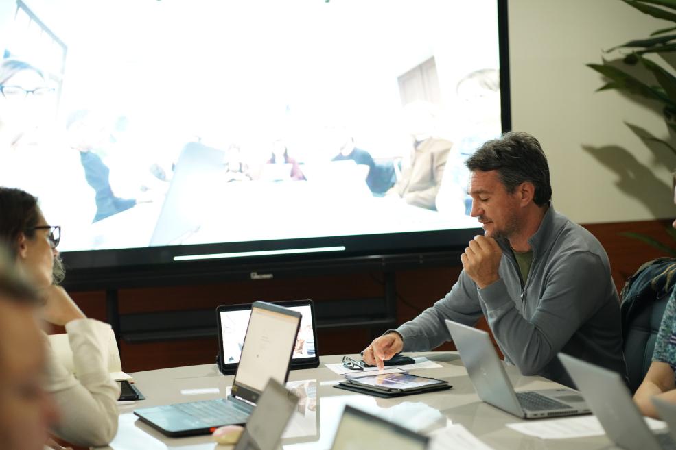 Several people sit at a table with laptops in a meeting room, with a large screen in the background showing an overexposed image.