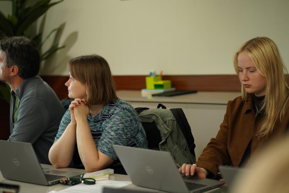 Three people sit at a table with laptops in a classroom or meeting room.