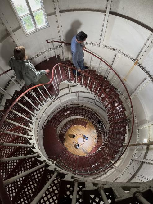 Top-down view of a spiral staircase with two people descending red metal steps inside a white tower with a small window.