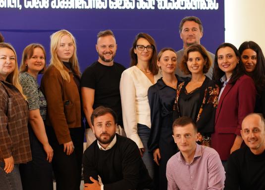 Group portrait of about fifteen people indoors in front of a large blue screen with white text at the top.