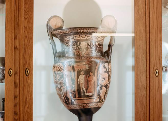 Liela antīka vāze ar sarkanfigūru gleznojumu eksponēta stikla vitrīnā muzeja interjerā.