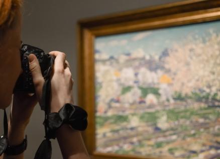 Sieviete ar fotoaparātu fotogrāfē gleznu, kurā attēlota pavasara ainava