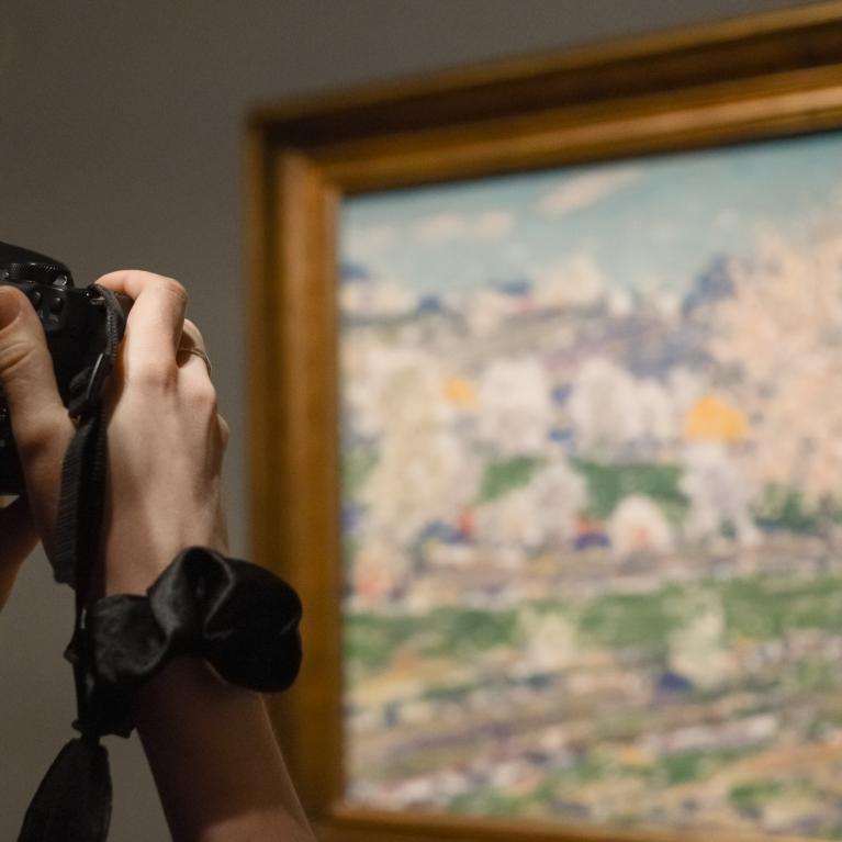 Sieviete ar fotoaparātu fotogrāfē gleznu, kurā attēlota pavasara ainava