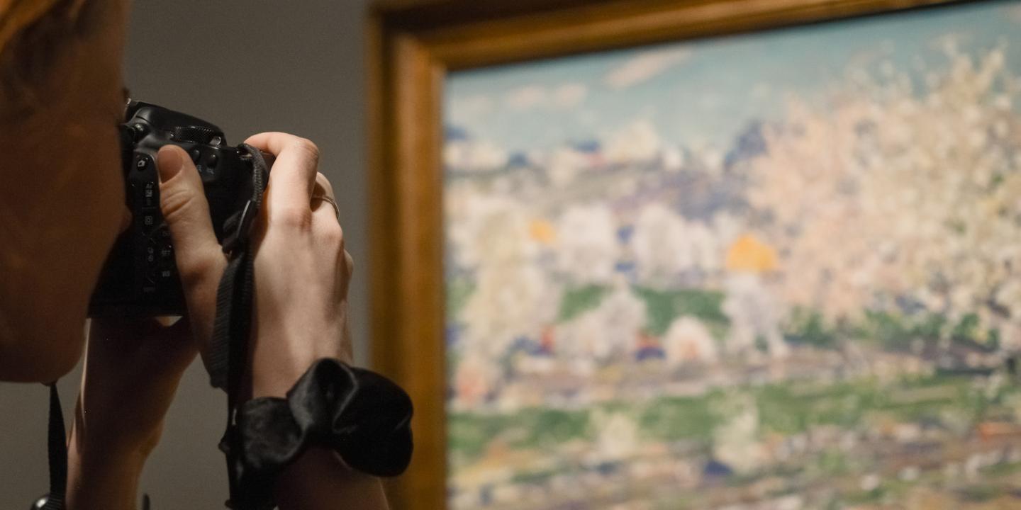 Sieviete ar fotoaparātu fotogrāfē gleznu, kurā attēlota pavasara ainava