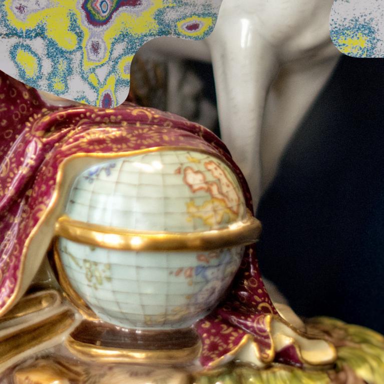 Tuvplānā redzama dekoratīva porcelāna figūra ar bordo un zeltītu auduma drapējumu, roku ar globusu un krāsainu fonu.