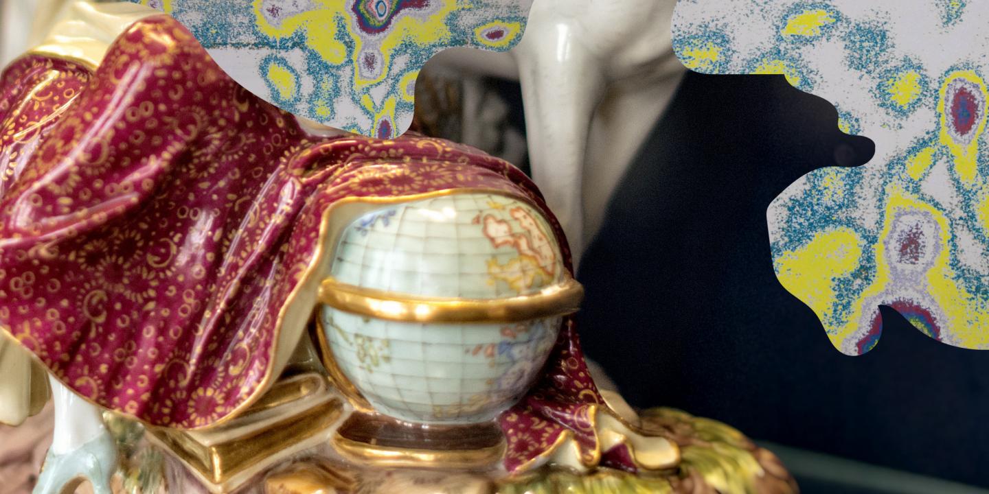 Tuvplānā redzama dekoratīva porcelāna figūra ar bordo un zeltītu auduma drapējumu, roku ar globusu un krāsainu fonu.