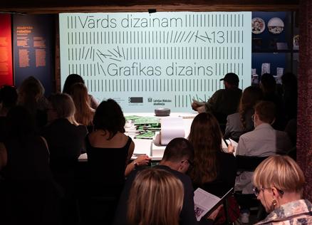 Muzeja zālē auditorija sēž skatītāju rindās pie izgaismota ekrāna ar uzrakstiem Vārds dizainam un Grafikas dizains, fonā redzami izstādes stendi.