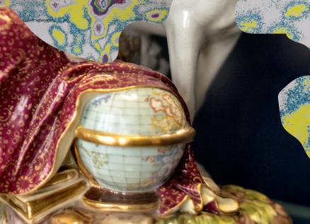 Tuvplānā redzama dekoratīva porcelāna figūra ar bordo un zeltītu auduma drapējumu, roku ar globusu un krāsainu fonu.