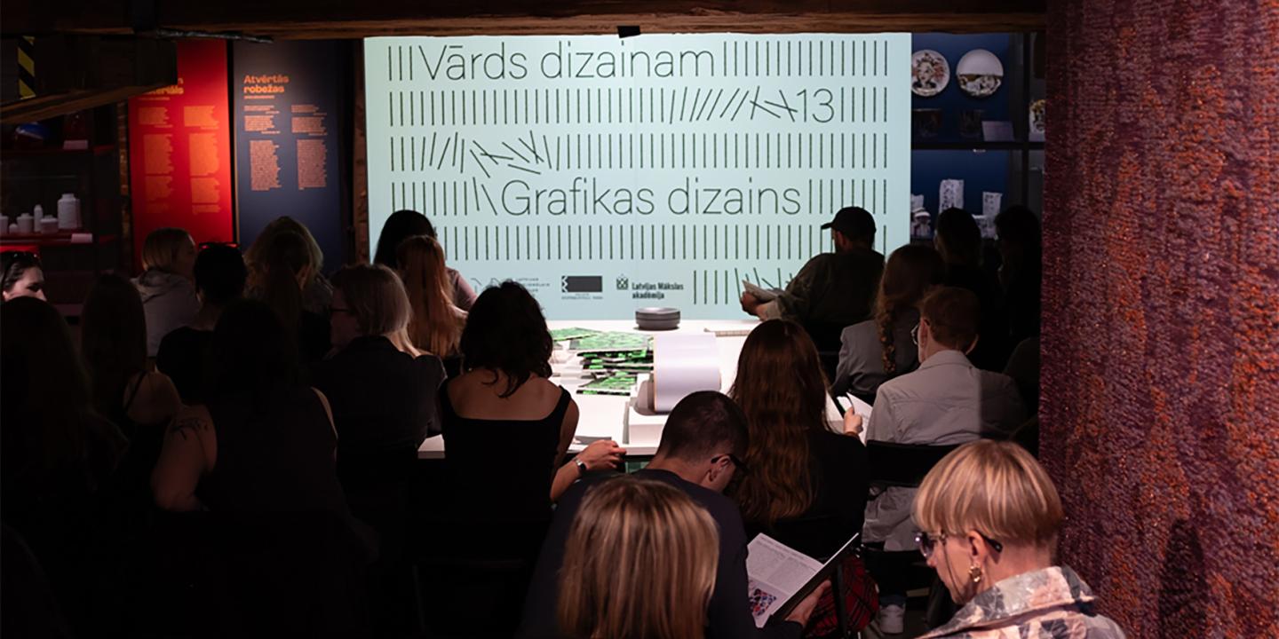 Muzeja zālē auditorija sēž skatītāju rindās pie izgaismota ekrāna ar uzrakstiem Vārds dizainam un Grafikas dizains, fonā redzami izstādes stendi.