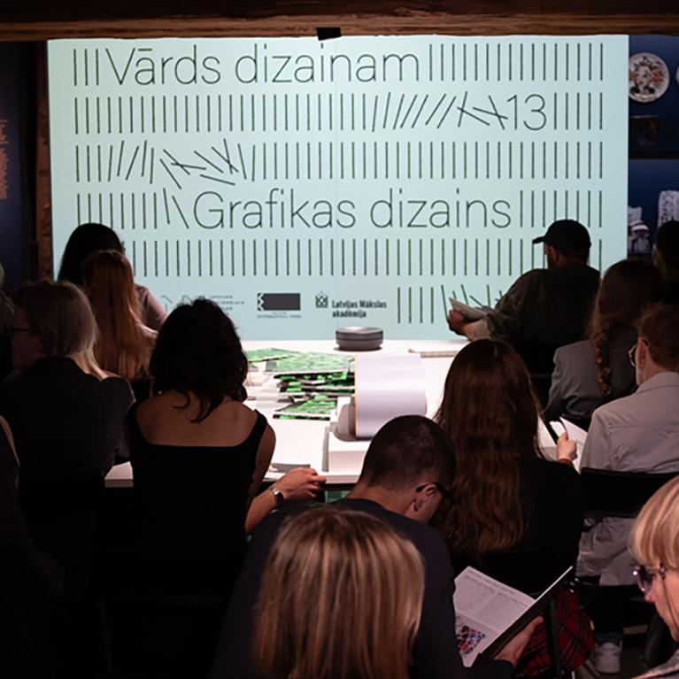 Muzeja zālē auditorija sēž skatītāju rindās pie izgaismota ekrāna ar uzrakstiem Vārds dizainam un Grafikas dizains, fonā redzami izstādes stendi.