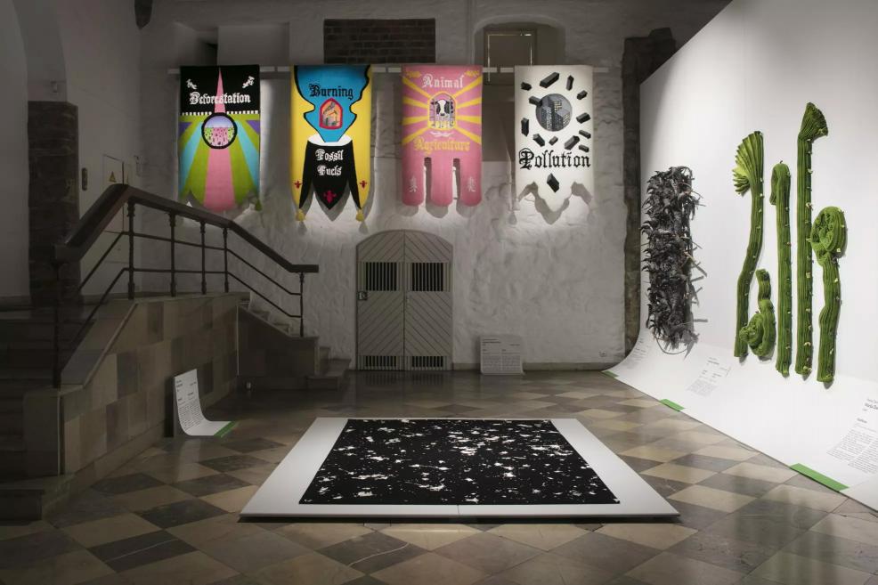Exhibition hall with banners on deforestation, fossil fuels, animal agriculture, and pollution, alongside textile and spatial artworks.