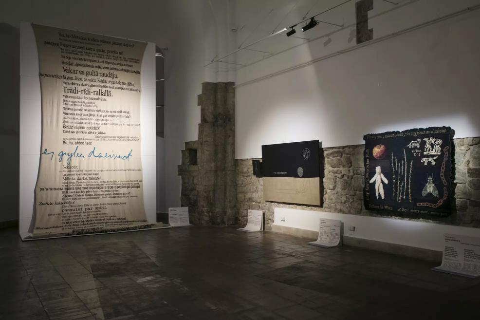 Exhibition hall with a large canvas featuring Latvian text and black and colorful textile works on the walls.