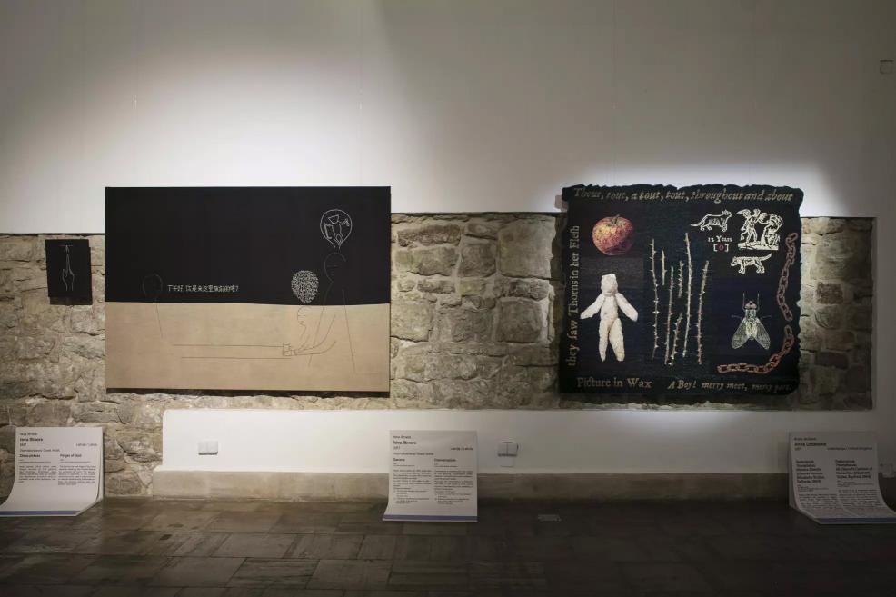 Two textile works on a stone wall – one black with linear figures and one dark with symbols, objects, and English inscriptions.
