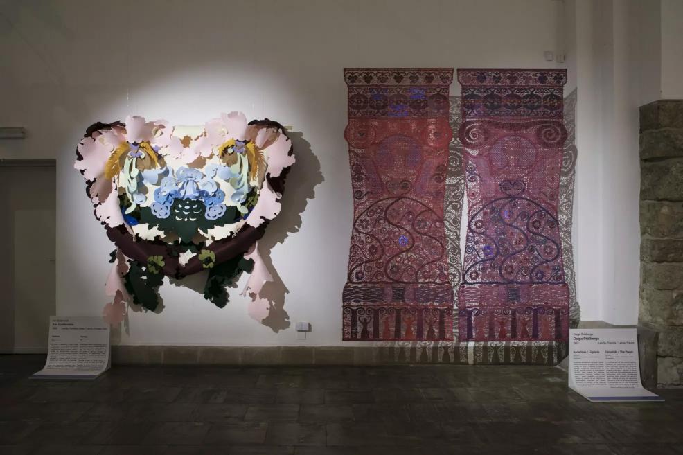 A large colorful three-dimensional floral composition and two ornamental red textile works displayed on the wall.