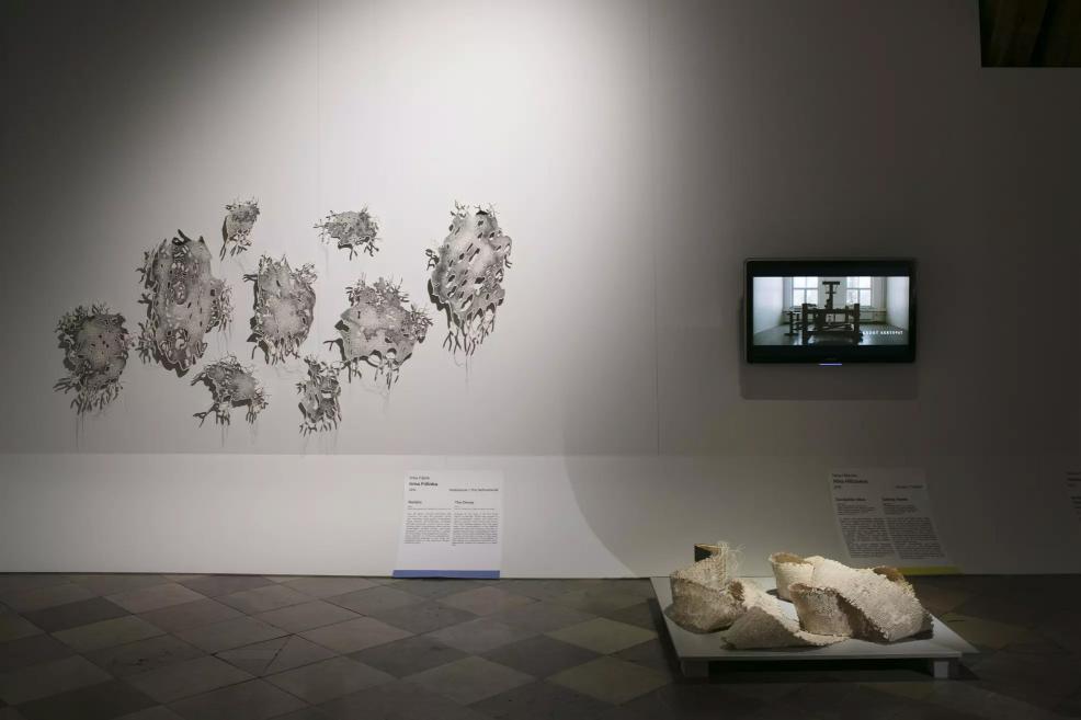 Exhibition hall with grey organic-shaped wall pieces, a video screen, and white sculptural objects on the floor.