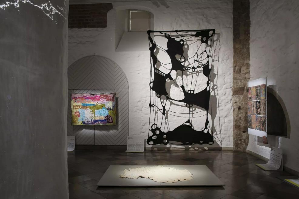 Exhibition hall with a black transparent textile installation, a colorful painting in the background, and a white sculptural work on the floor.