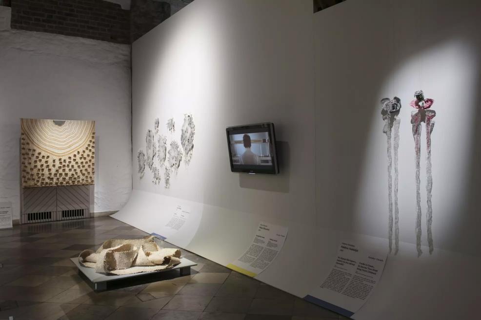 Exhibition hall with a large textile work of concentric lines, organic grey wall forms, and white sculptural objects on the floor.