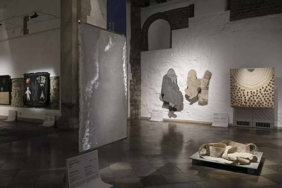 Exhibition hall with a semi-transparent fabric panel, textile works depicting human figures, and a geometric structure in the background.