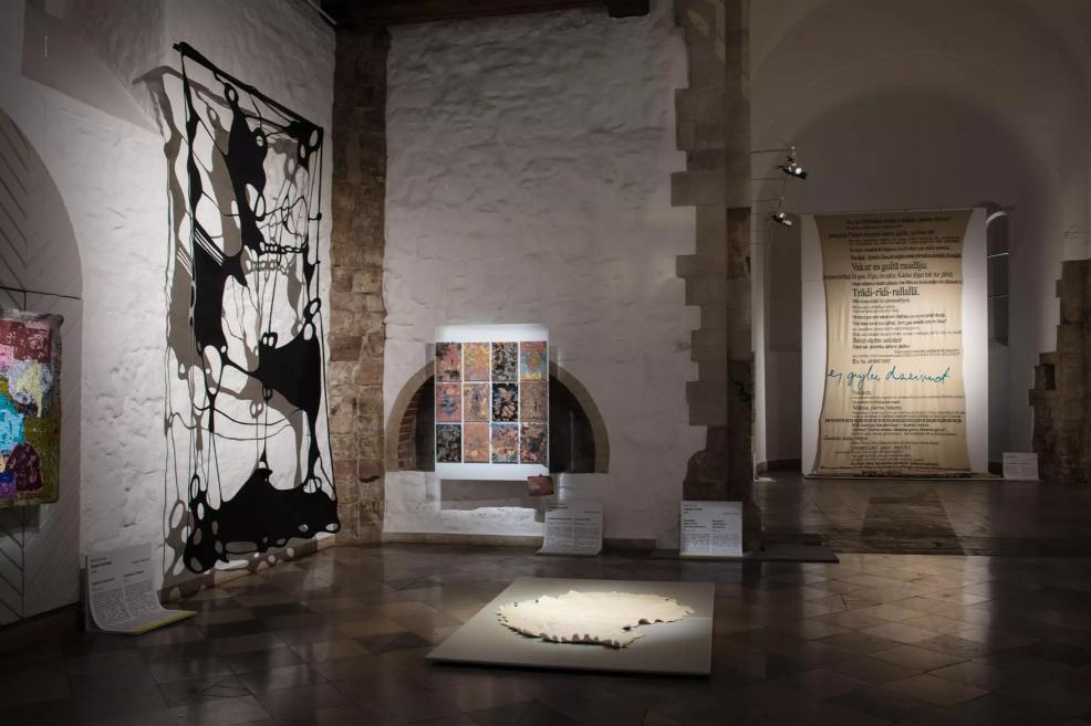 Exhibition hall with a black abstract textile installation, colorful panels, a large canvas with Latvian texts, and a white sculptural work on the floor.