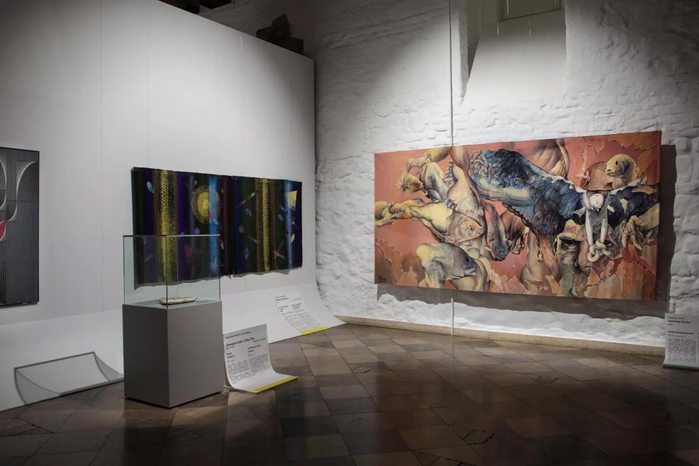 Exhibition hall with a dark ornamental textile piece and a large painting depicting human bodies, fish, and a bird.