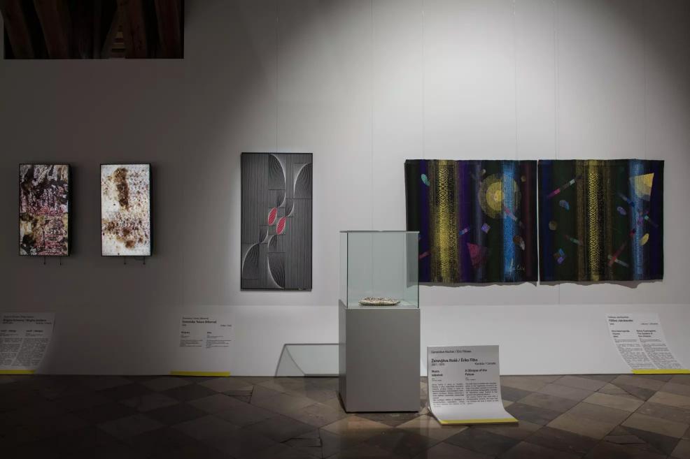 Three different artworks on the wall and a two-part dark textile work with geometric patterns, with a glass display case containing a small object in the foreground.