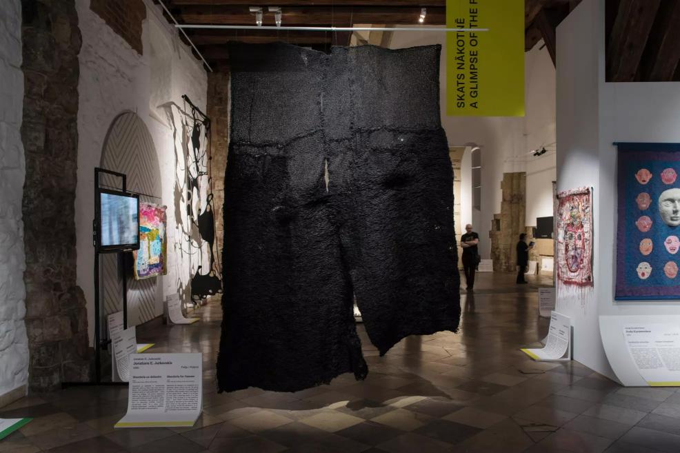 Exhibition hall with a large black textile installation in the center, colorful and figurative works on the walls, and a video screen in the background.