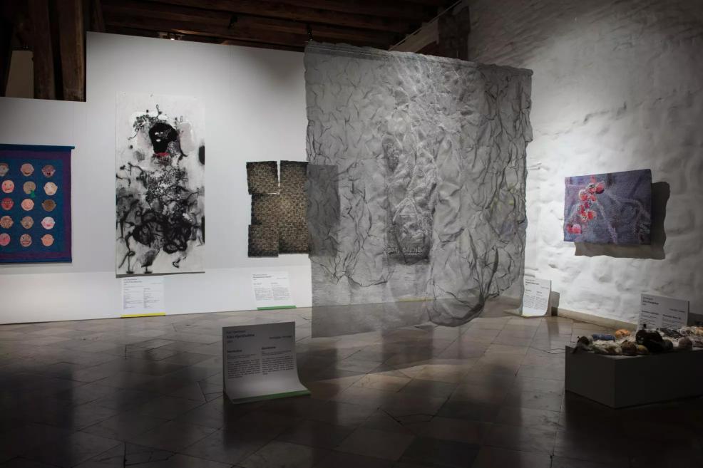 Exhibition hall with various textile works, including a grey translucent installation in the middle of the space.