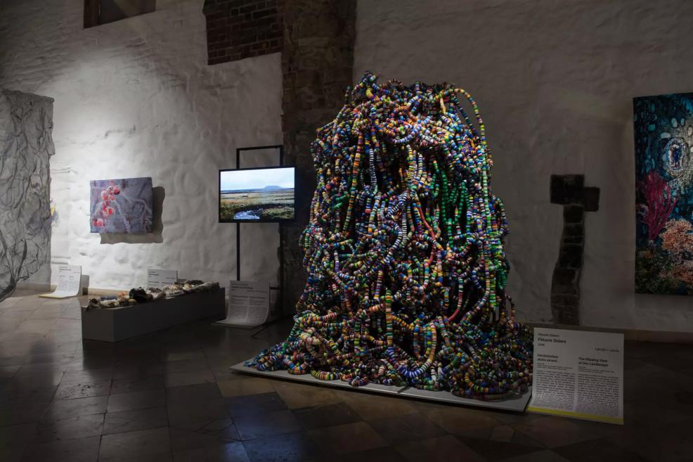 Large spatial installation made of colorful strands in the exhibition hall, with a video screen and other textile works in the background.