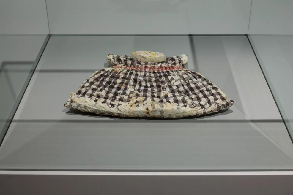 A small textile work of a checkered child’s dress displayed in a glass case.