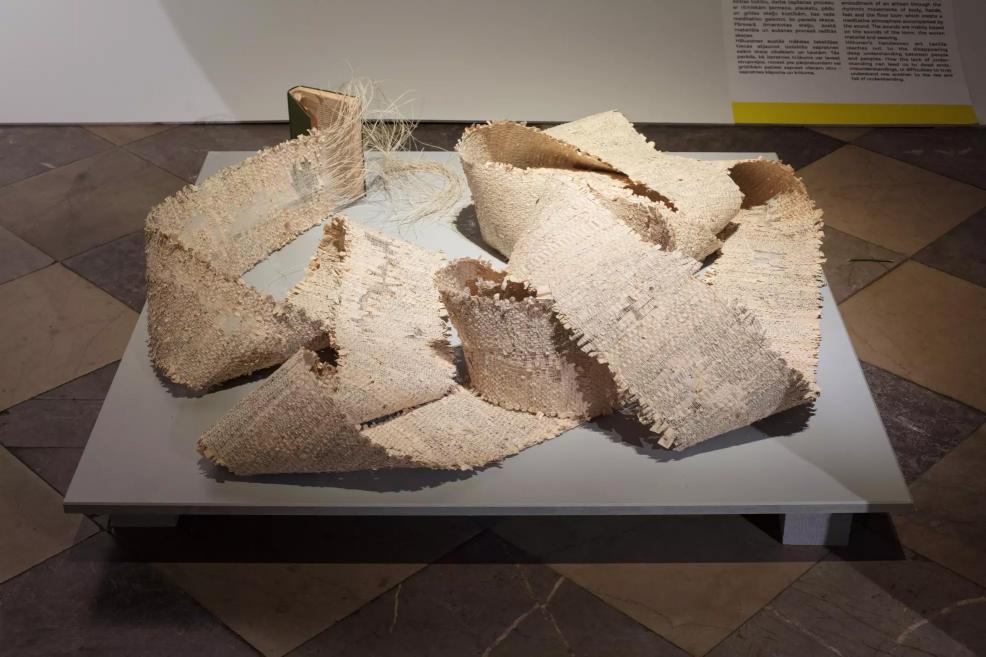 A light-colored textile work on a platform, made of twisted, ribbon-like structures.