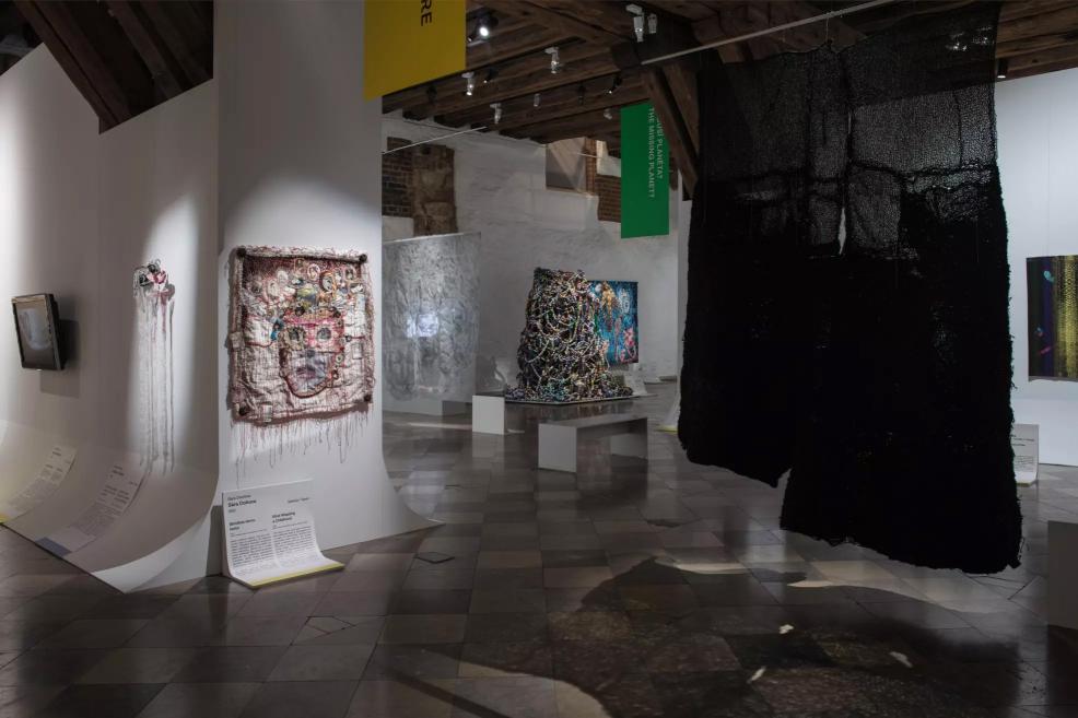 Exhibition hall with various contemporary textile art pieces, including a black textile installation, colorful fabrics, and spatial objects.