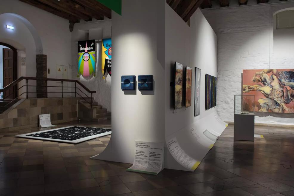 Exhibition hall with series of works along the walls, black fabric banners in the background, a dark floor installation, and a display case in the foreground.