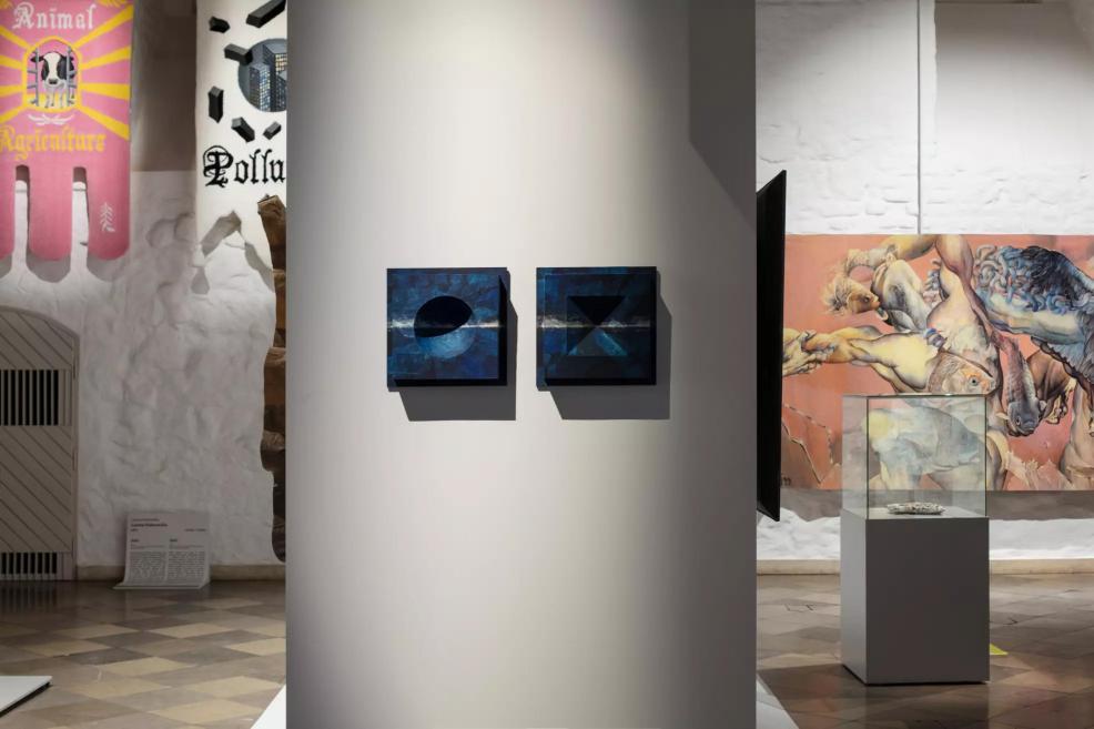 Two small square blue artworks displayed on a white stand, with tapestries and figurative fabrics visible in the background.