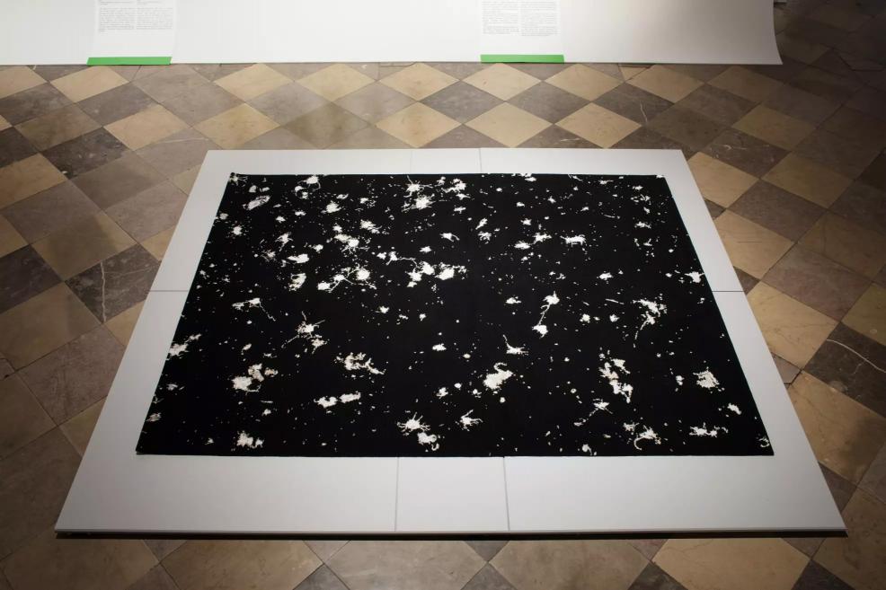 Black textile artwork with white splashes and speckled patterns displayed on the floor.