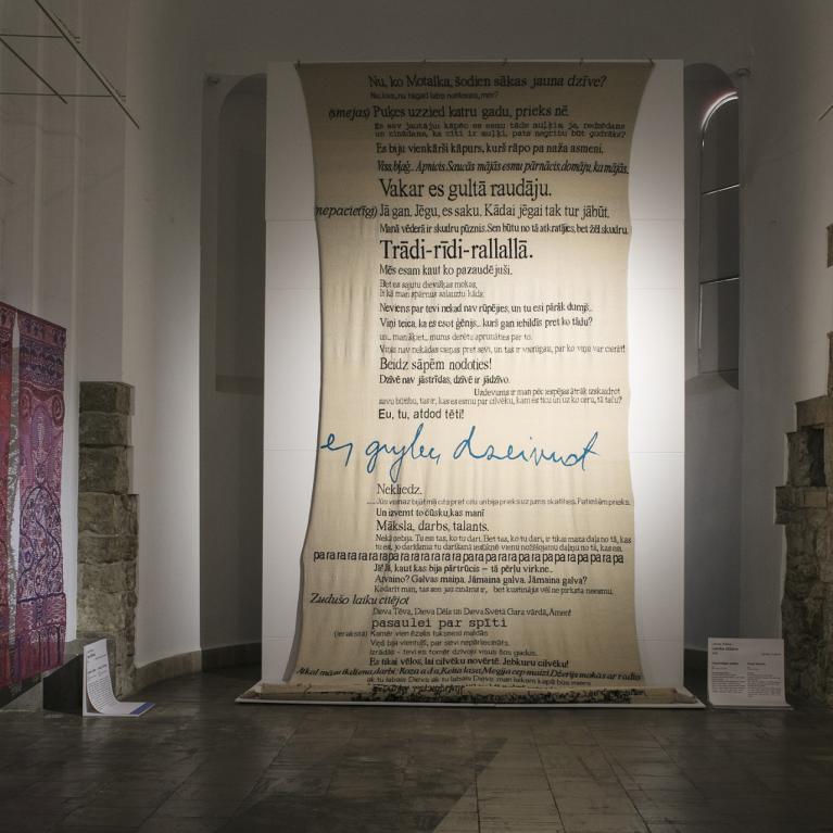 Contemporary art exhibition hall with a three-dimensional floral composition, textile works, a large canvas with inscriptions, and a black canvas with linear figures.