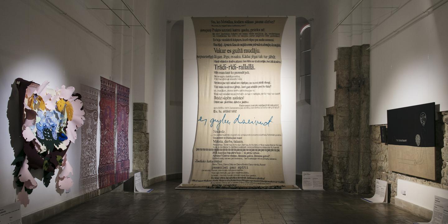 Contemporary art exhibition hall with a three-dimensional floral composition, textile works, a large canvas with inscriptions, and a black canvas with linear figures.