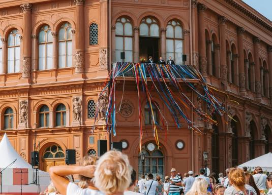 Rīgas Biržas ēkas balkons ar krāšņām daudzkrāsainām lentēm un cilvēku pūli laukumā svinīgā pasākumā.