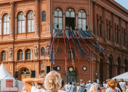 Rīgas Biržas ēkas balkons ar krāšņām daudzkrāsainām lentēm un cilvēku pūli laukumā svinīgā pasākumā.