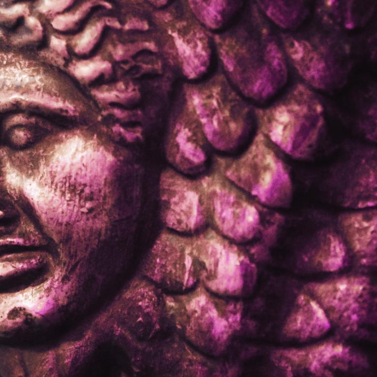 Bronzas reljefa fragments ar cilvēka seju un zvīņveida ornamentu violetā apgaismojumā.