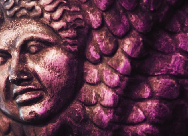 Bronzas reljefa fragments ar cilvēka seju un zvīņveida ornamentu violetā apgaismojumā.
