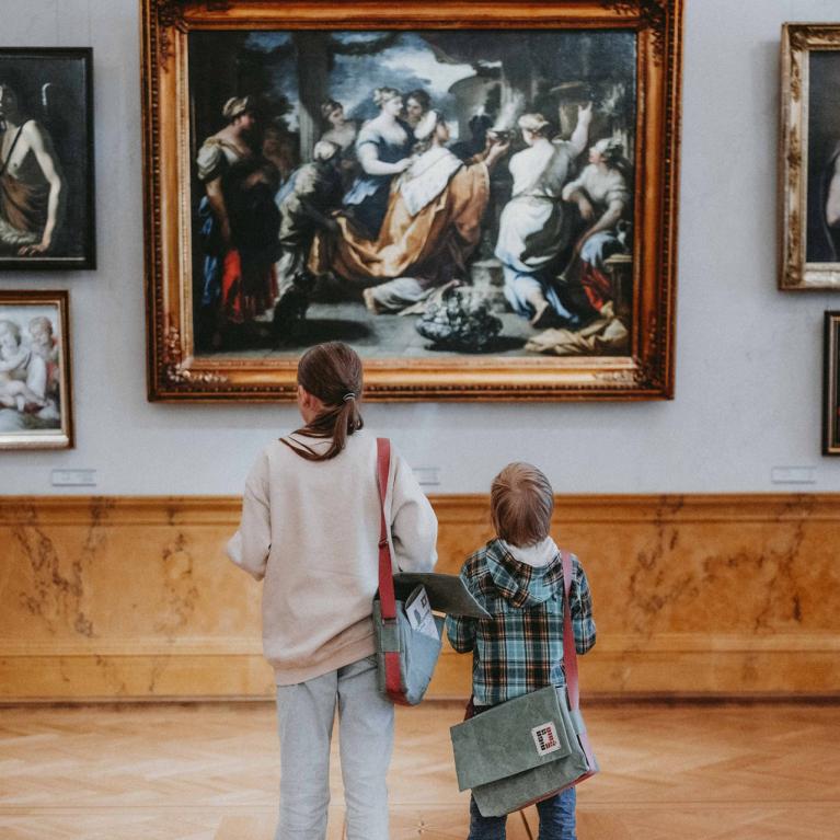 Divi bērni ar pleca somām muzeja zālē aplūko gleznas ar zeltītiem rāmjiem.