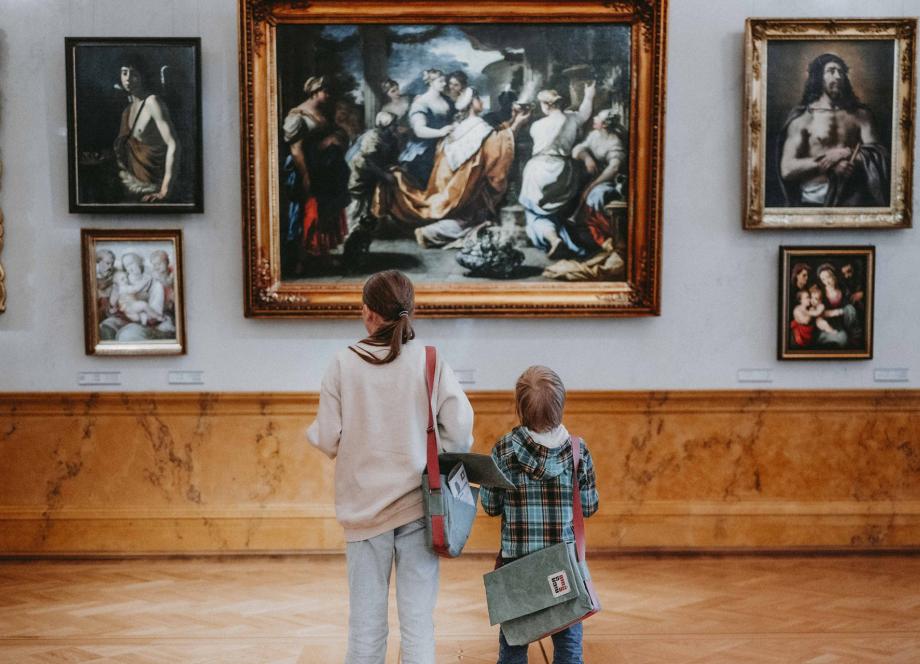 Divi bērni ar pleca somām muzeja zālē aplūko gleznas ar zeltītiem rāmjiem.