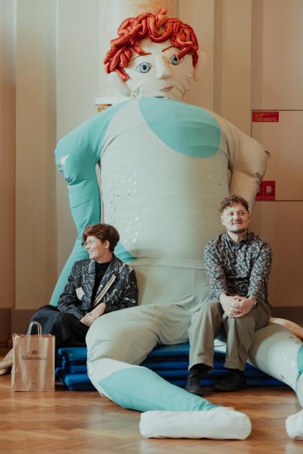 Two people sit in a gallery space beside a large soft sculpture with red hair and pale blue clothing.